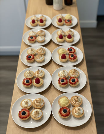 White plates lined on a wooden counter holding bite-sized fruit and lemon tarts topped with fresh berries and toasted meringue, arranged in neat rows for serving