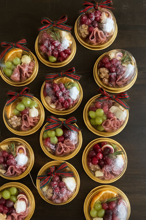 Overhead shot of mini holiday charcuterie cups on a dark wood table — clear dome lids tied with red-green plaid bows, each filled with grapes, sugared cranberries, an orange slice, cheese, cured-meat rosettes, walnuts and rosemary, styled as festive party favors.