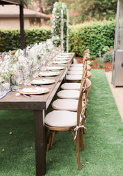 Long rustic wooden table set for an outdoor garden reception with white floral centerpieces, glassware and plates, and a row of tied cushioned wooden chairs on green lawn