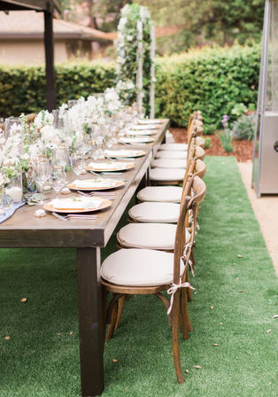 Long rustic wooden table set for an outdoor garden reception with white floral centerpieces, glassware and plates, and a row of tied cushioned wooden chairs on green lawn