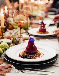 Poached red pear dessert topped with an edible purple flower and gold leaf, nestled on crunchy crumble and red sauce on a white plate at an elegant candlelit dinner table with wine glasses.