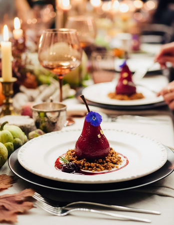 Poached red pear dessert topped with an edible purple flower and gold leaf, nestled on crunchy crumble and red sauce on a white plate at an elegant candlelit dinner table with wine glasses.