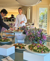 Catered gourmet buffet in a sunlit dining room — chef in a white coat arranging colorful salads and appetizers on a glass table with purple floral centerpieces and a balcony palm-tree view.