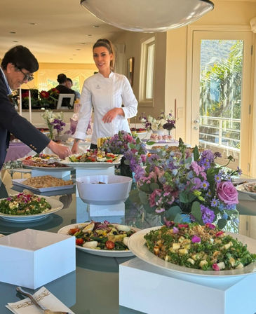 Catered gourmet buffet in a sunlit dining room — chef in a white coat arranging colorful salads and appetizers on a glass table with purple floral centerpieces and a balcony palm-tree view.