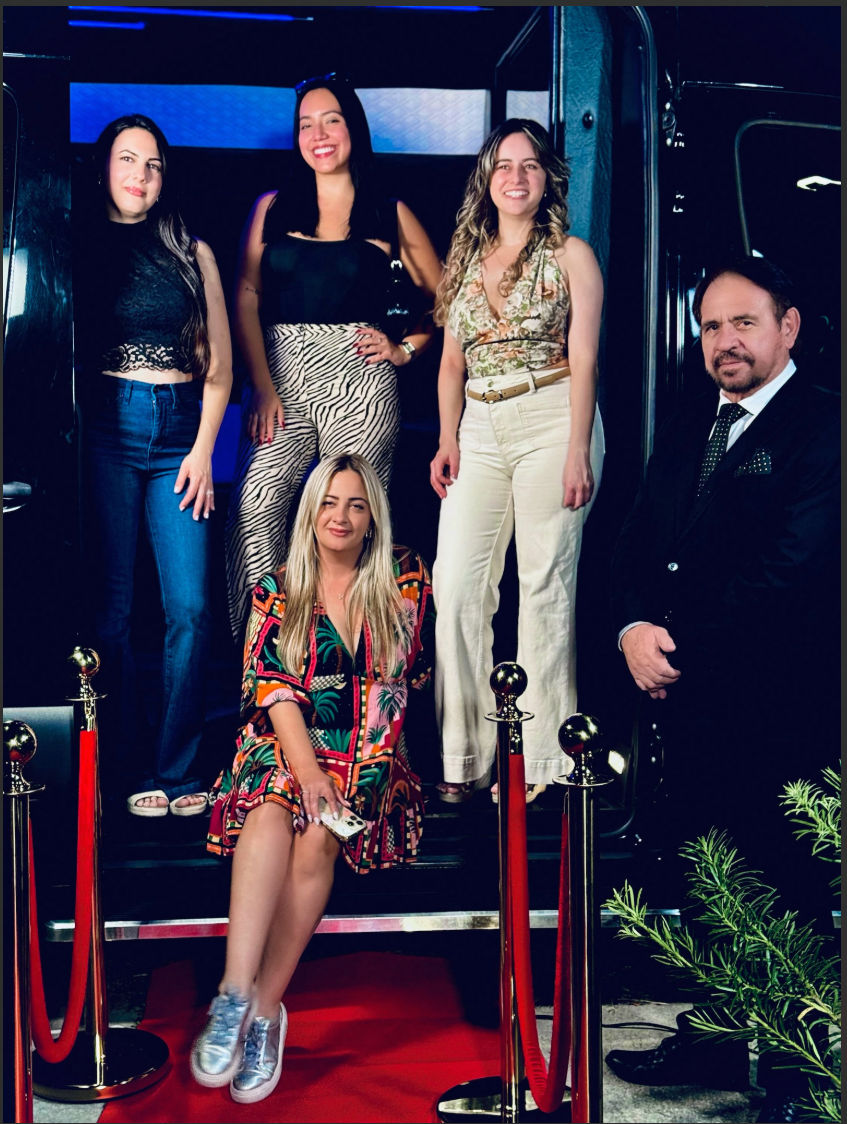 Five people posing on red-carpet steps framed by gold stanchions and velvet ropes — a woman in a colorful dress and silver sneakers seated in front, three women in casual-chic outfits standing behind, and a suited man at the side, giving a fun night-out/event vibe.