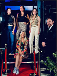 Five people posing on red-carpet steps framed by gold stanchions and velvet ropes — a woman in a colorful dress and silver sneakers seated in front, three women in casual-chic outfits standing behind, and a suited man at the side, giving a fun night-out/event vibe.