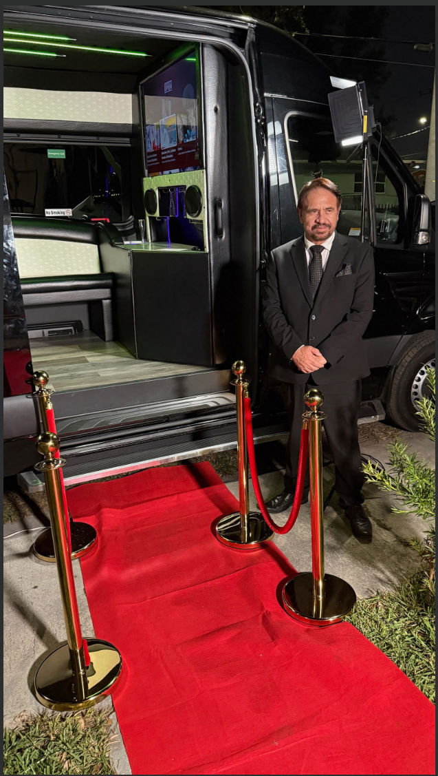 Well-dressed man in a black suit stands beside an open luxury party van at night, with a red carpet and gold stanchions leading into a neon-lit interior with screens.