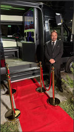 Well-dressed man in a black suit stands beside an open luxury party van at night, with a red carpet and gold stanchions leading into a neon-lit interior with screens.