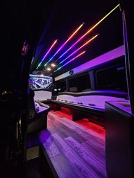 Luxury party bus interior at night with rainbow LED ceiling strips, wraparound leather bench seating, wood-look flooring and ambient colored floor lighting