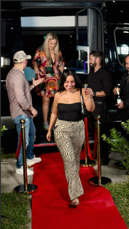 Cheerful woman in zebra-print pants and a black strapless top walks down an outdoor evening red carpet from a party bus, holding a champagne flute as other guests chat behind velvet ropes.