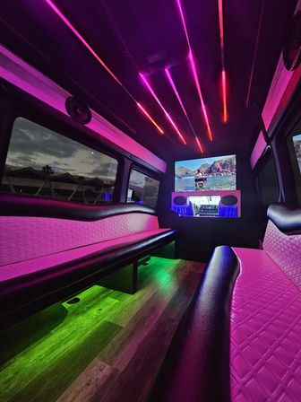 Luxury party van interior with neon pink and purple LED ceiling strips, green under-seat floor lighting, quilted pink leather bench seating, wood-look flooring and a wall-mounted flat-screen TV playing a beach scene.