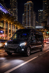 Sleek black Mercedes van cruising Brickell at night — palm trees wrapped in lights and towering Miami skyscrapers glowing in the background.