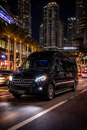 Sleek black Mercedes van cruising Brickell at night — palm trees wrapped in lights and towering Miami skyscrapers glowing in the background.