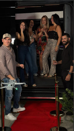 Friends laughing and toasting with champagne on the steps of a party bus at a red carpet entrance, with stanchions and ropes - lively nightlife celebration.