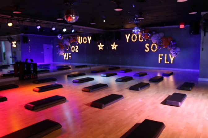 Bright indoor fitness studio set for a step workout — rows of black step platforms on a wooden floor, mirrored wall, disco balls and purple-orange lighting with marquee bulbs reading 'YOU SO FLY'.