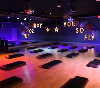 Bright indoor fitness studio set for a step workout — rows of black step platforms on a wooden floor, mirrored wall, disco balls and purple-orange lighting with marquee bulbs reading 'YOU SO FLY'.