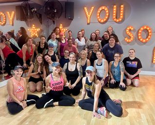 Smiling group of about 30 women in athletic wear posing after a group fitness class in a bright studio with hardwood floor, mirrored wall and marquee light letters and stars.