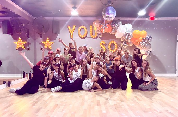 Energetic group fitness class photo in a mirrored dance studio — smiling participants pose on a hardwood floor beneath marquee letters, illuminated star lights, disco balls and a metallic balloon arch for a celebratory workout event.