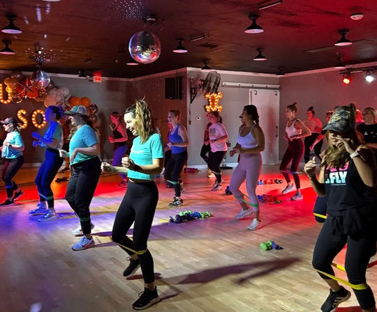 Energetic group fitness class in an indoor studio with disco ball and colorful lights — participants doing a cardio-dance workout with resistance bands and hand weights.