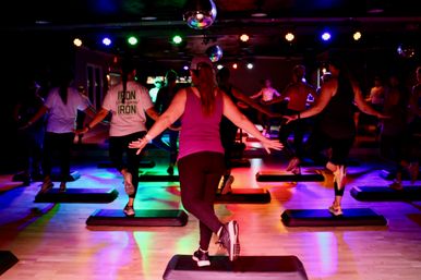 Energetic group step aerobics class in an indoor fitness studio with disco balls, colorful stage lights, and participants on aerobic step platforms.