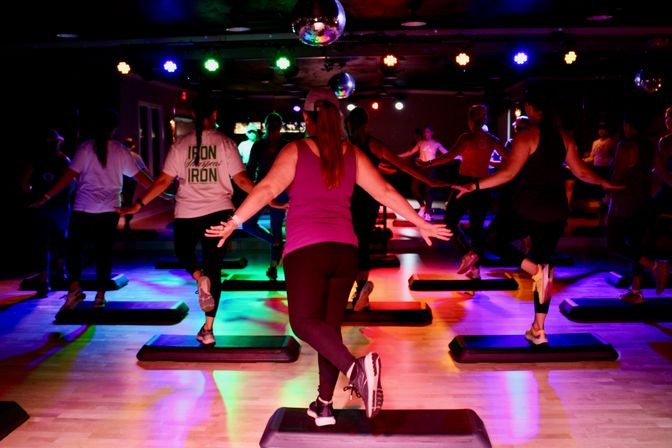 Energetic group step aerobics class in an indoor fitness studio with disco balls, colorful stage lights, and participants on aerobic step platforms.