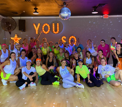 Large group of women in neon workout clothes smiling and posing on a wood floor in a dance studio under a disco ball and lit marquee letters reading "YOU" after a disco-themed group fitness class