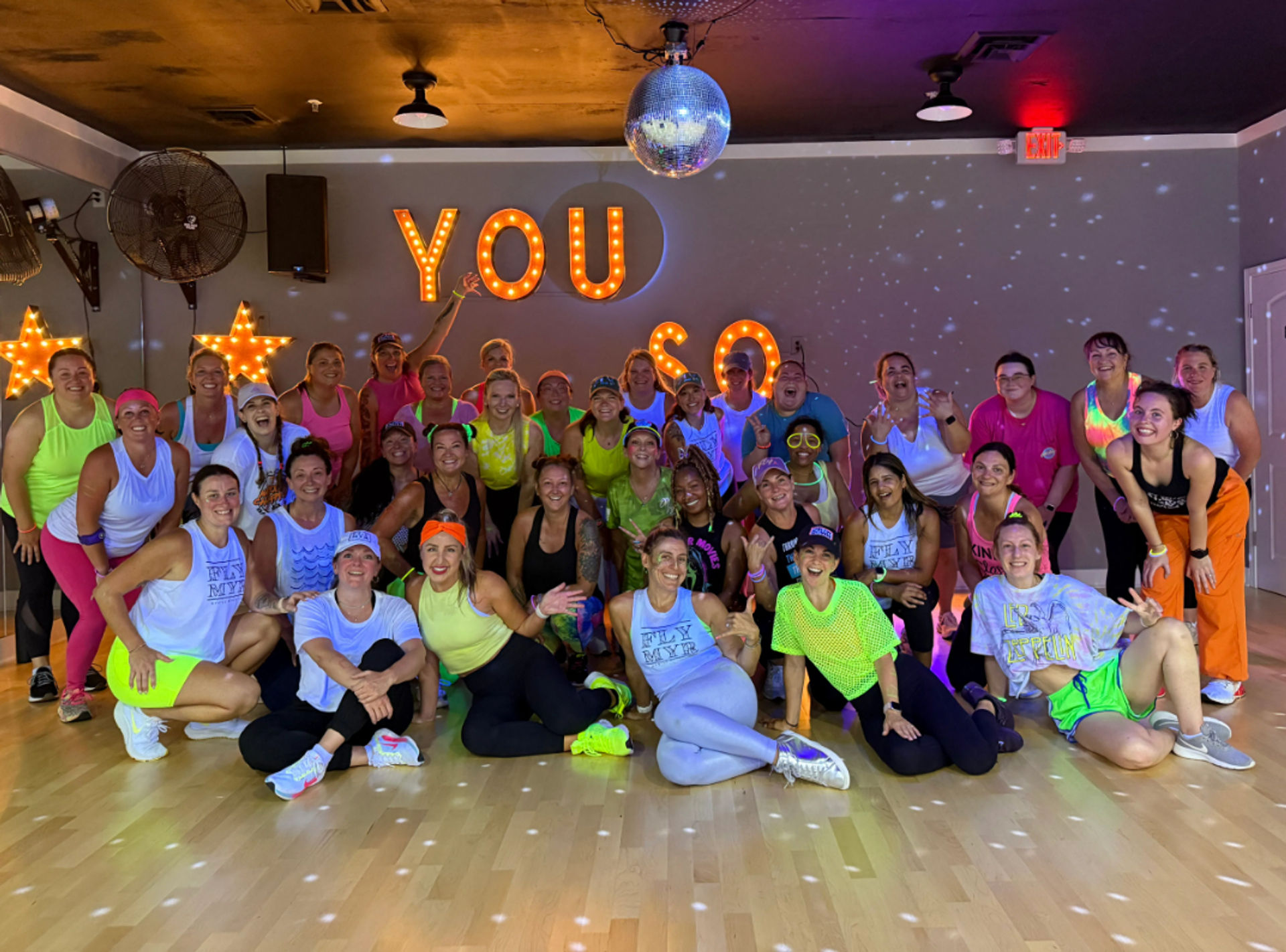 Large group of women in neon workout clothes smiling and posing on a wood floor in a dance studio under a disco ball and lit marquee letters reading "YOU" after a disco-themed group fitness class