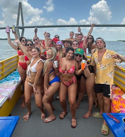 Group of friends in colorful swimwear partying on a yellow boat, holding drinks and smiling with a cable-stayed bridge and blue sky over a coastal river in the background.
