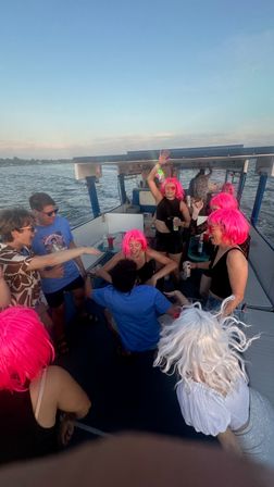Group of people wearing neon pink wigs dancing and laughing on a party boat at sunset with drinks and open water in the background
