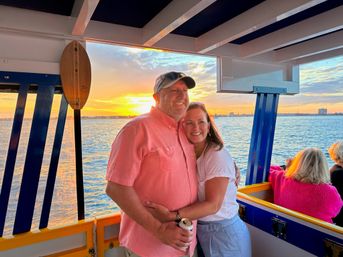 Smiling couple hugging on a colorful boat during a golden sunset cruise, holding a canned drink with calm blue water and a distant shoreline skyline.