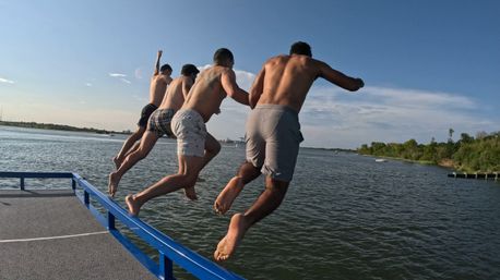 Four shirtless men jumping off a blue dock railing into a calm river on a sunny summer day, shoreline trees visible in the distance.