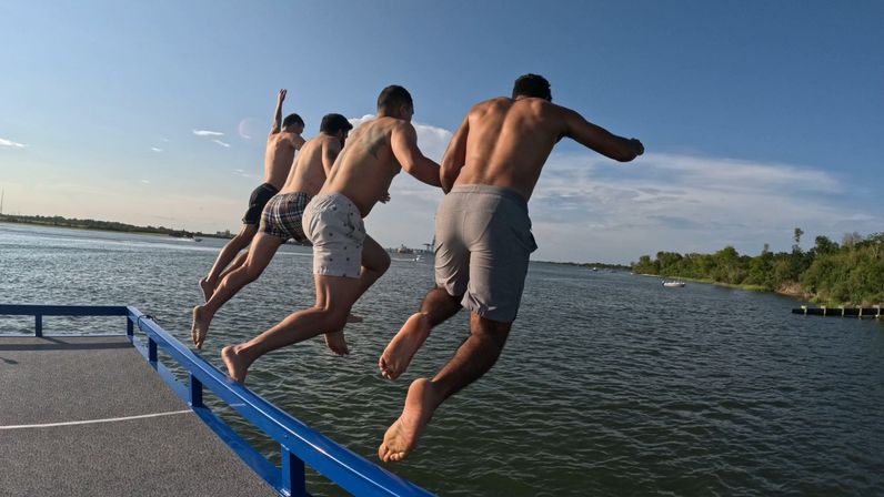 Four shirtless men jumping off a blue dock railing into a calm river on a sunny summer day, shoreline trees visible in the distance.