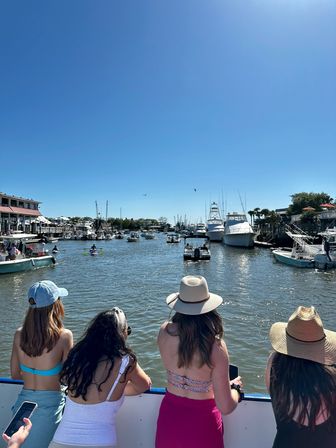 Four people in swimsuits and sun hats leaning on a boat railing, looking out at a busy coastal marina with boats, yachts and waterfront buildings under a clear blue sky