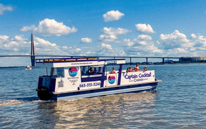 Party cruise boat with passengers cruising across a sunny coastal harbor, cable-stayed bridge in the background, blue sky with puffy clouds and shimmering water reflections.