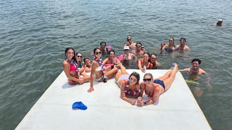 Large group of friends relaxing on a floating dock in a lake, wearing swimsuits and red-white-blue leis, smiling and holding drinks on a sunny summer day with a blue cap on the dock.