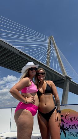 Two women in sunglasses and bikinis—one wearing a white sun hat with a veil—pose on a boat cruise deck beneath a dramatic cable-stayed bridge on a sunny blue day with a colorful tropical cocktail sign visible.