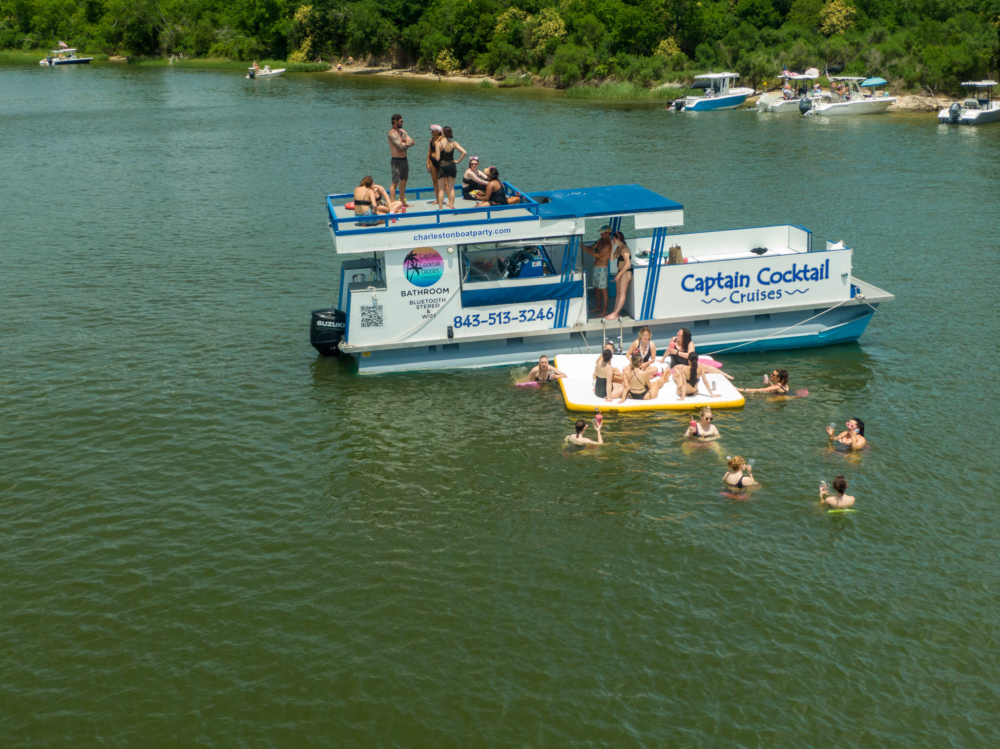 Party boat with people on deck and on a large inflatable platform, swimmers enjoying a sunny day in a tree-lined inlet.