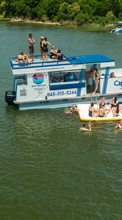 Party boat with people on deck and on a large inflatable platform, swimmers enjoying a sunny day in a tree-lined inlet.