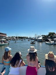 Four people in swimsuits and sun hats leaning on a boat railing, overlooking a sunny coastal marina filled with yachts, small boats and waterfront docks