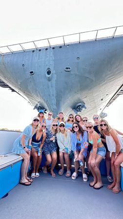 Group of friends in summer outfits smiling and posing on a small boat beneath the towering gray bow of a ship in a sunny coastal harbor, drinks in hand — summer boat outing.