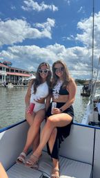Two friends in swimsuits and sunglasses, wearing leis and holding canned drinks, sitting on a boat at a sunny waterfront marina under a blue sky with fluffy clouds.
