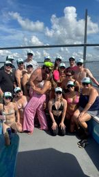 Smiling group of friends on a sunny boat near a cable-stayed bridge, many wearing matching green-and-white caps, swimsuits and sunglasses, with a couple kissing in the center under a blue sky with puffy clouds.