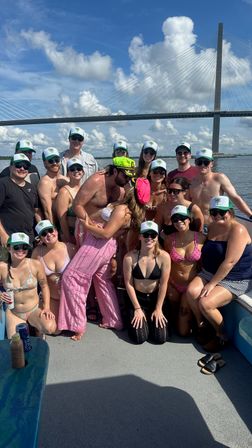 Smiling group of friends on a sunny boat near a cable-stayed bridge, many wearing matching green-and-white caps, swimsuits and sunglasses, with a couple kissing in the center under a blue sky with puffy clouds.