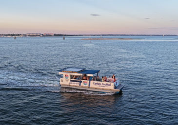 Small sightseeing cruise boat with passengers gliding across a calm coastal bay at sunset, blue water and distant shoreline