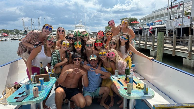 Large group of friends on a party boat at a marina, wearing colorful novelty sunglasses and laughing around painted tables with drinks, docks, yachts, and a waterfront boardwalk in the background.
