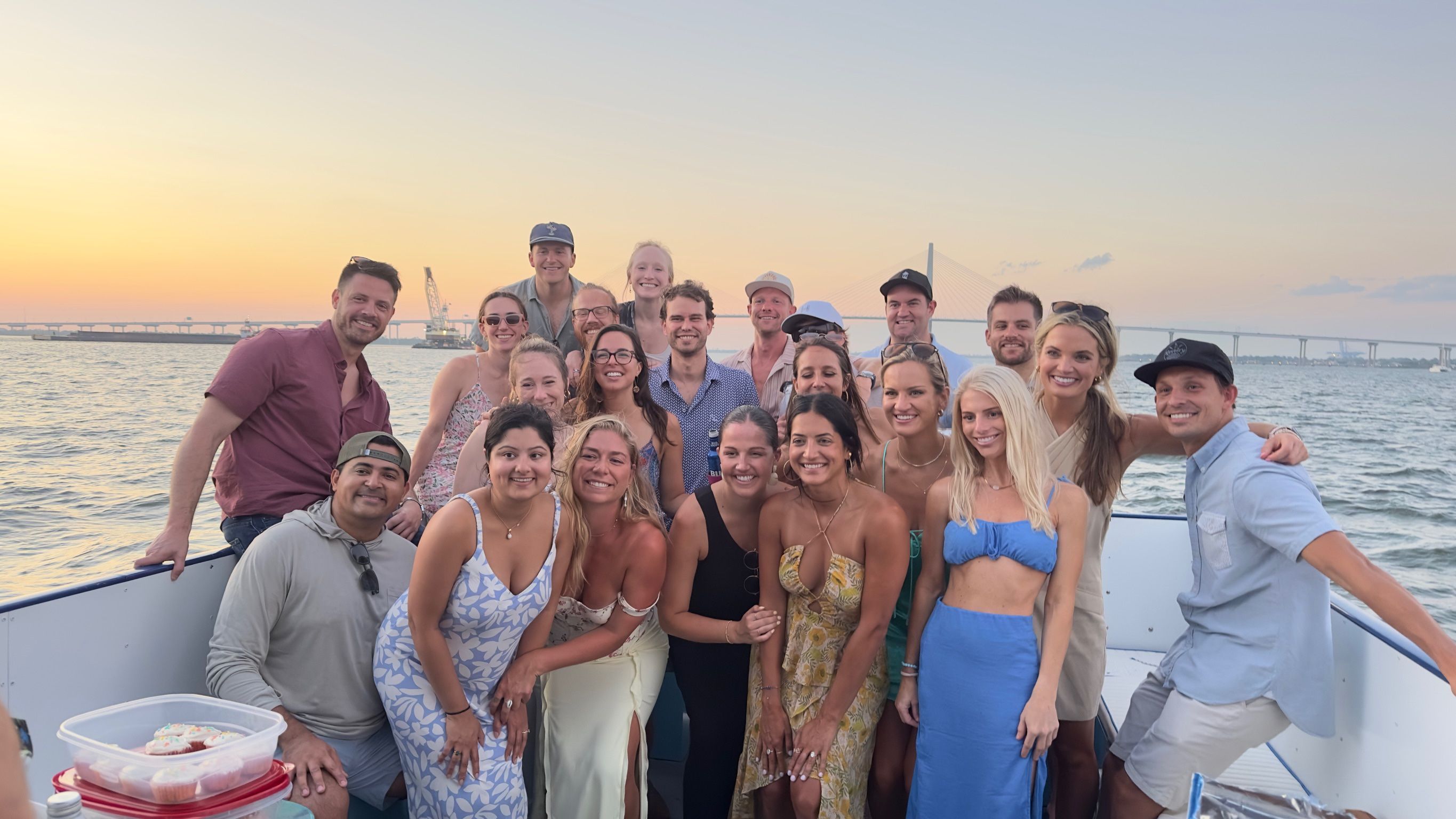 Group of friends smiling on a sunset boat cruise, wearing summer outfits with a suspension bridge and calm waterfront in the background