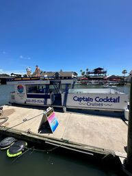 Party cruise boat moored at a sunny coastal marina with people on the upper deck, palm trees and waterfront restaurants in the background under a clear blue sky.