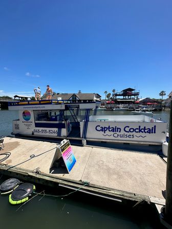 Party cruise boat moored at a sunny coastal marina with people on the upper deck, palm trees and waterfront restaurants in the background under a clear blue sky.