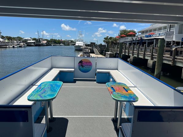 Sunlit open-top boat deck with white bench seating and two vibrant marbled tables, docked at a coastal marina with boats, wooden piers, palm trees and blue sky.