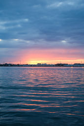 Coastal harbor sunset with a pink-orange horizon under blue clouds, rippling water reflecting warm light and a silhouetted shoreline with distant harbor lights.
